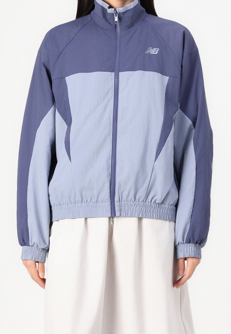 Žena oblečená v dvoubarevném svetle a tmavě modrém bomberu se zipem, s elastickými manžetami a lemem, spárovaná s bílou sukní.