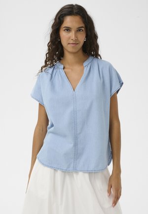 Femme aux longs cheveux bouclés portant un chemisier bleu clair à manches courtes et une jupe blanche, debout devant un fond uni.