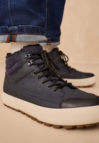 Sneakers alte navy con colletto in tessuto strutturato, lacci neri e suola in gomma color crema; cuciture dettagliate e motivo del battistrada robusto.