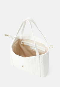 Hvit håndveske med et teksturert bladmønster, gullfarget detaljer, to håndtak og et beige interiør som har en glidelåslomme og en flat lomme.