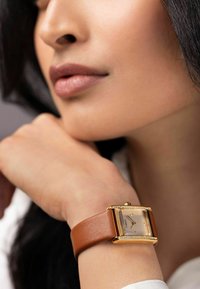 Montre en or rectangulaire avec un cadran métallique, bracelet en cuir marron et accents en cristal sur la lunette. Les détails incluent des repères horaires et une couronne.