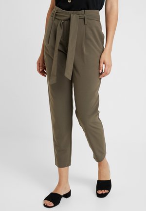 Femme portant un pantalon à taille nouée olive ajusté aux chevilles et des sandales à talons blocs noirs à bout ouvert, debout devant un fond uni.