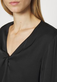 Blusa nera con scollatura a V, realizzata in tessuto liscio, caratterizzata da morbide pieghe sul petto e delicate cuciture lungo le cuciture.