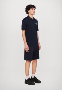 Mornarska polo majica s ovratnikom in vezenim logotipom, kombinirana s kratkimi hlačami v usklajeni barvi, ki imajo teksturiran stranski del, in črnimi športnimi čevlji.