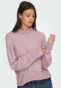 Kvinne iført en lilla genser med perledetaljer på høy hals og mansjettene, kombinert med blå jeans, ser ned og justerer ermene.