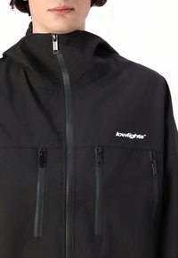 Veste légère noire avec capuche, dotée de deux poches zippées et d'un logo sur la poitrine. Texture lisse, design minimaliste.