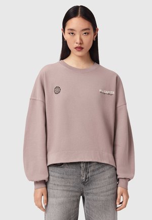Junge Frau mit langen dunklen Haaren trägt ein übergroßes mauvefarbenes Sweatshirt mit Globus- und "ALLSAINTS"-Logos, kombiniert mit grauen Jeans.