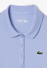 Polo de corte slim en azul claro, con tela texturizada, cierre de tres botones y un logo de cocodrilo verde en el lado izquierdo del pecho.