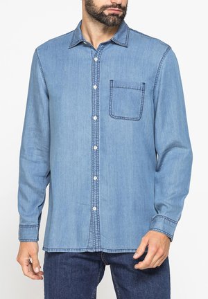 Uomo che indossa una camicia di jeans azzurro chiaro con bottoni, maniche lunghe e taschino sul petto, abbinata a jeans blu scuro.