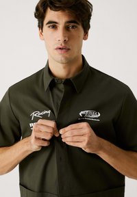 Camisa de botón de manga corta de color verde oscuro con acentos de logotipo, una textura suave y un cuello. Manos abrochando los botones delanteros.