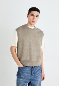 Pull en tricot sans manches beige avec un col rond, texture douce et ourlet côtelé, porté par-dessus une chemise blanche à manches courtes et un jean bleu.