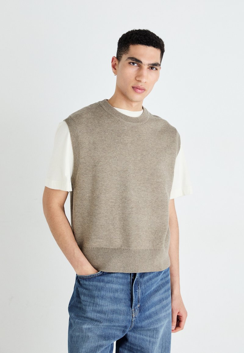Pull en tricot sans manches beige avec un col rond, texture douce et ourlet côtelé, porté par-dessus une chemise blanche à manches courtes et un jean bleu.