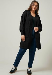 Schwarze Strickjacke mit einem strukturierten Rautenmuster, offener Vorderseite und langen Ärmeln, kombiniert mit dunkelblauen Jeans und schwarzen Sneakern.