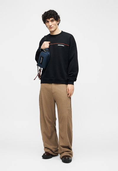 Jonge man met een zwarte DSQUARED2 trui, beige cargobroek, bruine schoenen en een marineblauwe crossbody tas tegen een effen witte achtergrond.