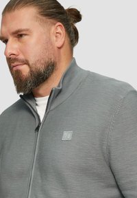 Grauer Reißverschluss-Pullover mit strukturiertem Strickmuster und geripptem Kragen, ausgestattet mit einem kleinen Logo auf der linken Brust.