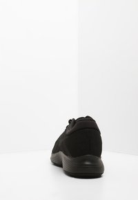 Zapatilla deportiva negra con parte superior de malla texturada, collar acolchado y suela de goma. Presenta cordones y un diseño aerodinámico.