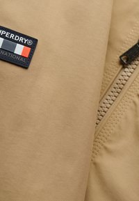 Tan jakke af stof med ribbet tekstur, med en lynlås og en sort logo-patch med røde, hvide og blå accenter.