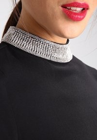 Svart topp med en hög, utsmyckad krage av metalliska rhinestones som reflekterar ljus. Tyget ser slätt och kroppsnära ut.