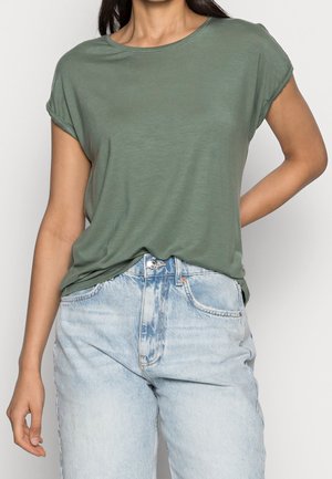 Woman wearing a muted green short-sleeve t-shirt tucked into light blue high-waisted jeans with hand on hip.