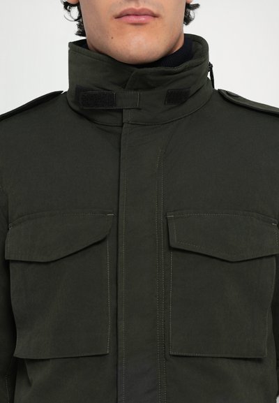 Dunkelgrüne Jacke mit hohem Kragen, Fronttaschen und Klettverschluss. Hergestellt aus strukturiertem Stoff mit sichtbaren Nähdetails.