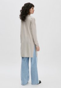 Lang, lichtbeige cardigan met zijslitsen, gedragen over lichtblauwe flared jeans. De stof lijkt zacht en licht van gewicht; het heeft lange mouwen.