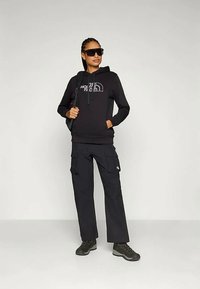 Kvinna klädd i svart hoodie, cargobyxor, vandringsskor, ryggsäck och stora solglasögon, stående mot en enfärgad grå bakgrund.