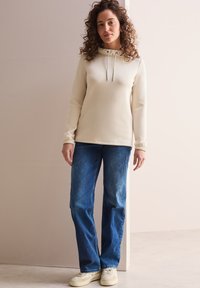 Kremowy sweter z kapturem i sznurkiem, w połączeniu z niebieskimi prostymi dżinsami i jasnymi trampkami. Widoczne kręcone włosy.