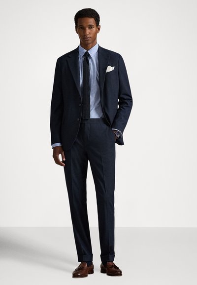 Vīrietis tērpies tumši zilu uzvalkā ar gaiši zilu krekls, melnu adītu kaklasaiti, baltu kabatas lakatiņu, brūnām kurpēm ar bārkstīm un rokas pulksteni.