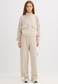 Conjunto deportivo beige con chaqueta corta con cremallera y dos bolsillos frontales, y pantalones de pierna ancha con bolsillos laterales tipo cargo. Textura suave.