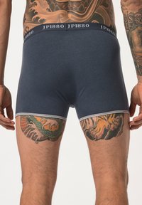 Námořnické modré boxerky s kontrastním bílým lemem a elastickým pasem s logem "JP1880"; model ukazuje složité tetování na stehnech.