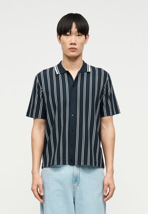 Jeune homme portant une chemise noire à manches courtes avec des rayures blanches verticales et un jean en denim bleu clair, debout devant un fond uni.