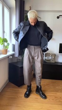 Person med blekt hår som bär svart läderjacka, marinblå tröja, beige plisserade byxor och svarta stövlar, stående inomhus vid fönstret.