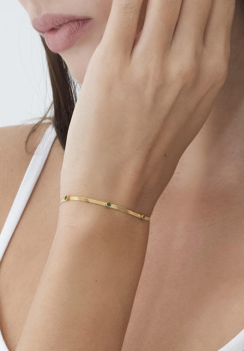 Brățară de aur cu un design subțire, având accente de pietre verzi și o textură netedă, purtată pe un încheietură cu un ton de piele neutru.