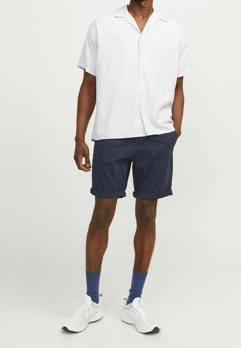 Homme portant une chemise blanche à manches courtes boutonnée, un short bleu marine à pois retroussé, des chaussettes bleues et des baskets blanches, debout les mains détendues.
