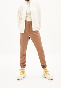 Giacca beige a zip sopra una maglia a coste chiara, abbinata a joggers beige a gamba affusolata e stivali bianchi con lacci gialli. Texture del tessuto liscia.
