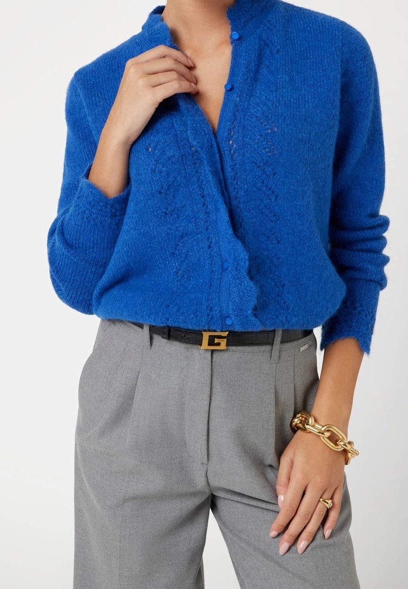 Femme portant un cardigan tricoté bleu avec des bords festonnés, un pantalon gris taille haute, une ceinture noire avec boucle dorée, et un bracelet chaîne en or.