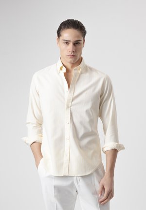 Joven con el cabello peinado hacia atrás, vestido con una camisa de botones amarilla clara con las mangas remangadas y pantalones blancos, posando frente a un fondo liso.