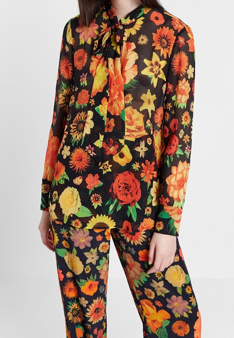 Personne portant un chemisier à manches longues et un pantalon avec un imprimé floral coloré orange, jaune et rouge sur fond noir, debout.