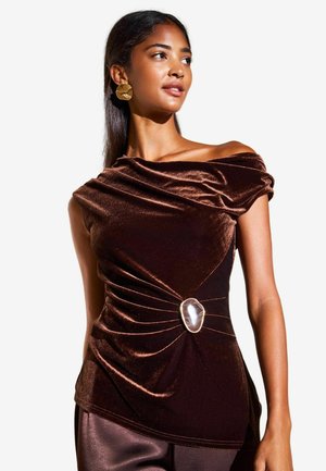 Jonge vrouw draagt een donkerbruine fluwelen off-shoulder top met een decoratieve zilveren broche en grote gouden oorbellen.