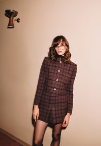 Giacca fitted a quadri marroni con bottoni dorati, colletto alto e gonna mini abbinata. La modella indossa occhiali, con uno sfondo di parete liscia e apparecchio di illuminazione.