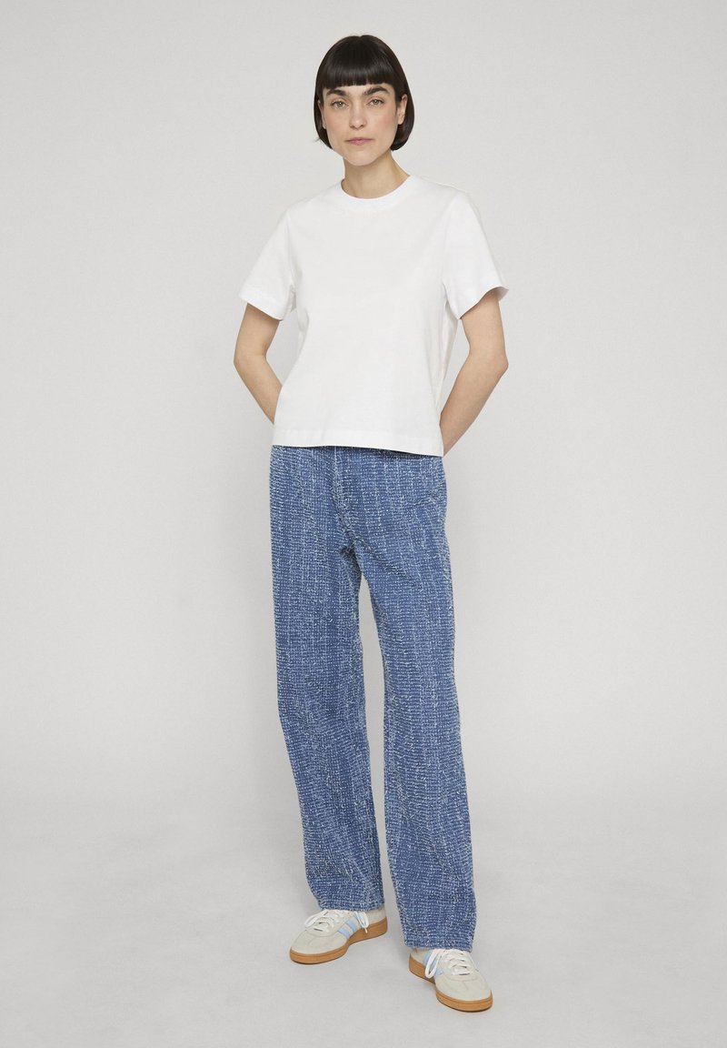 Νεαρή γυναίκα στέκεται με τα χέρια πίσω από την πλάτη, φοράει λευκό κοντομάνικο πουκάμισο, μπλε υφασμάτινο παντελόνι με φαρδιά μπατζάκια και μπεζ αθλητικά παπούτσια.