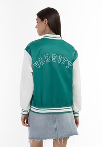 Zelená a bílá baseballová bunda s vyšitým nápisem "VARSITY" na zadní straně, s pruhovanými manžetami a lemem, vyrobená z hladkého, lesklého materiálu.