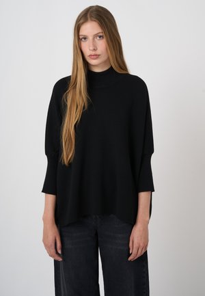 Young woman with long blonde hair wearing a loose black long-sleeve top and dark pants, standing against a plain light background.