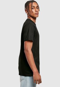 Black cotton t-shirt with a classic crew neck, short sleeves, and a relaxed fit. Model shown wearing light blue jeans, side view.