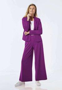 Blazer ajusté violet et pantalon large, fabriqués à partir d'un tissu lisse. Associé à un T-shirt blanc et des baskets blanches.