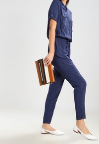 Marinblå jumpsuit med korta ärmar och två framfickor, i kombination med vita ballerina-skor och en rektangulär brun och krämfärgad clutch.