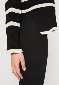 Pull à rayures noires et blanches avec un motif tricoté texturé, présentant un ourlet côtelé contrastant et une coupe décontractée, associé à un pantalon noir.