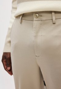 Beige housut, joissa on sileä tekstuuri, napin suljenta ja sivutaskut. Kangas vaikuttaa kevyeltä ja hengittäviltä.