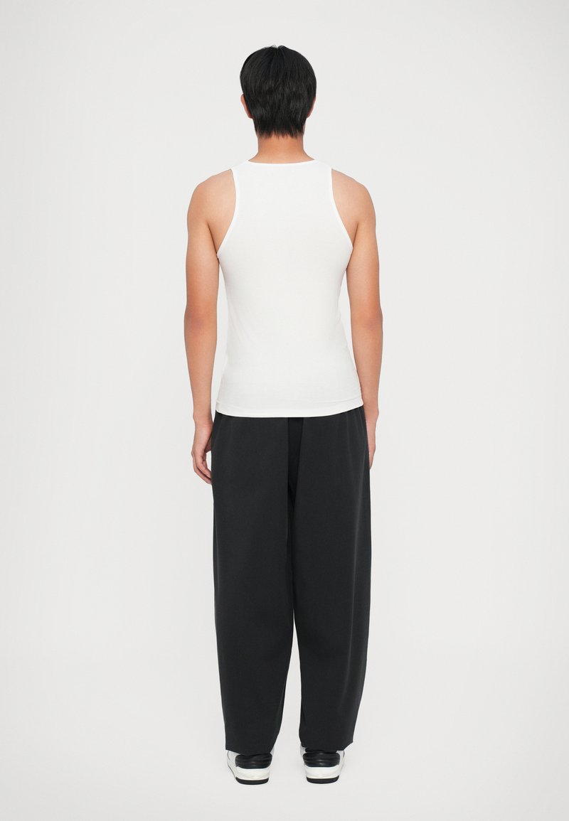 Hvid ærmeløs tanktop med ribbet tekstur, parret med løstsiddende sorte bukser. Model vist fra ryggen, iført sorte sneakers.