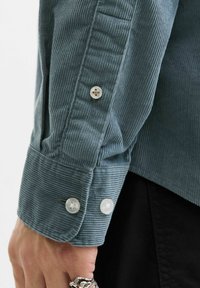 Blå kordfløyelsmansjett med hvite knapper og subtil ribbestruktur, som viser en knappdetalj og en skreddersydd kant nederst.